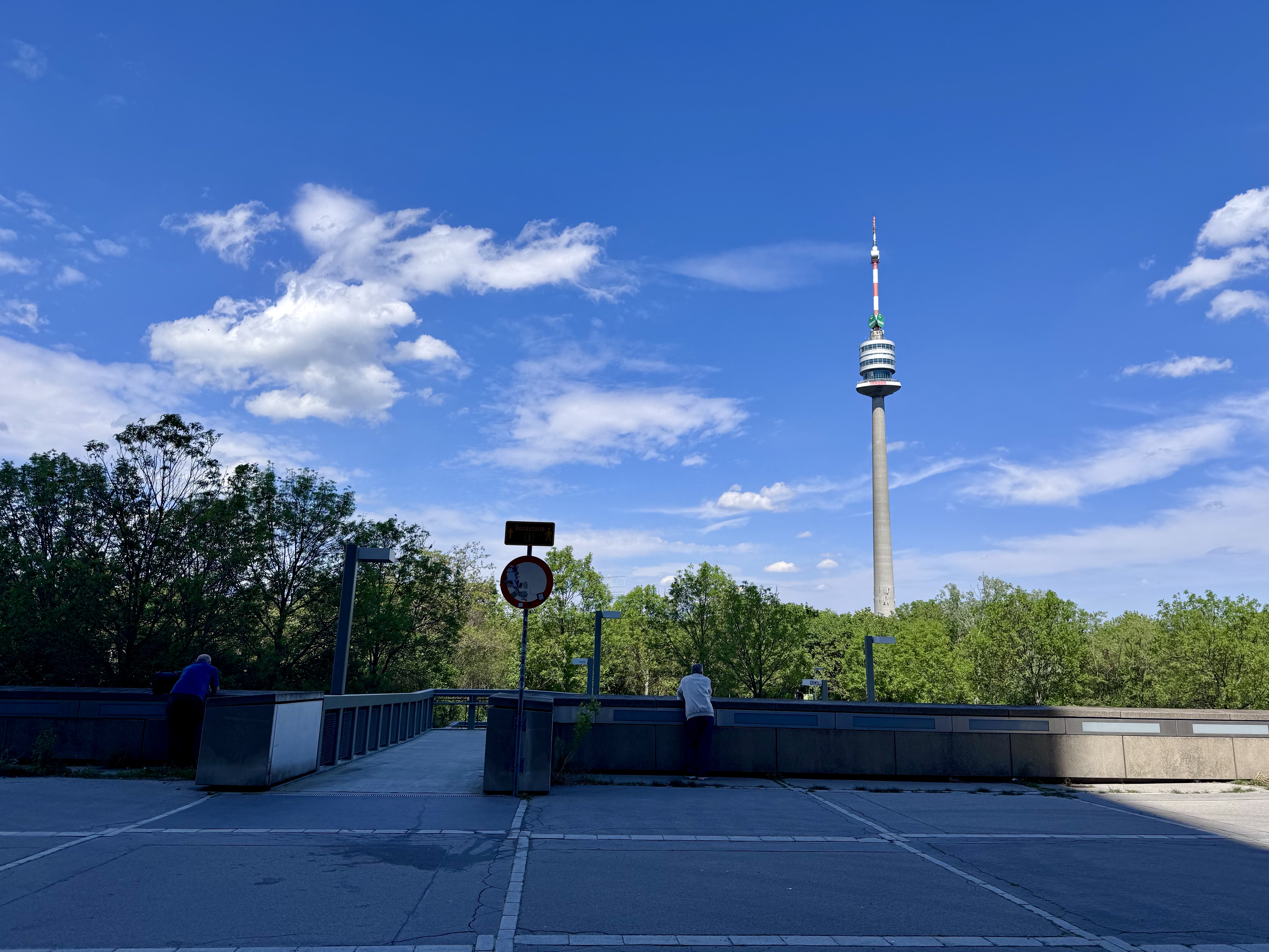 A view of the blue sky with a few scattered clouds, a tower, and trees beyond a compound wall